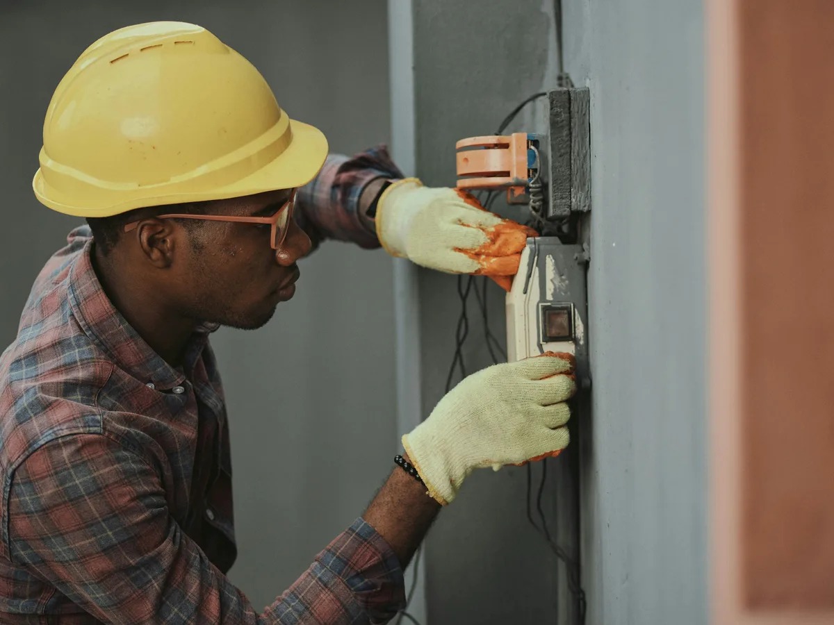 An electrician in a hard hat and gloves working on a meter box